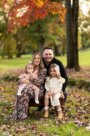 A family sits together on grass surrounded by vibrant autumn foliage and fallen leaves in a park setting.