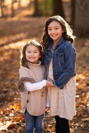 Two children share a sweet embrace while standing on fallen autumn leaves in the woods wearing cozy sweaters and denim.