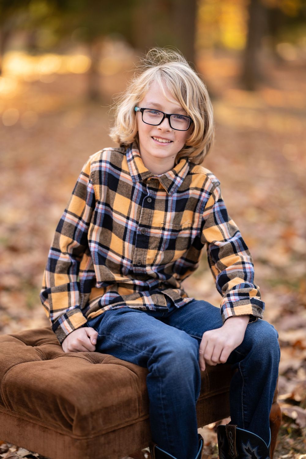 Child in plaid flannel shirt and jeans sits on a brown velvet cushion surrounded by autumn leaves on the ground.