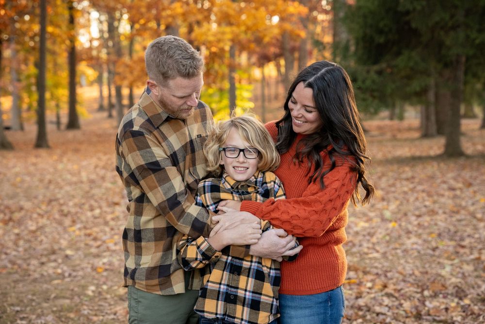 A couple embraces outdoors in a park with autumn leaves and golden light filtering through trees in the background.