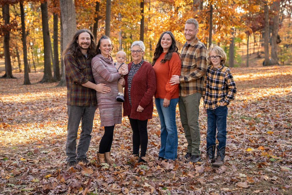 A family group stands together smiling on fallen autumn leaves in a sunlit wooded area on a crisp fall day.