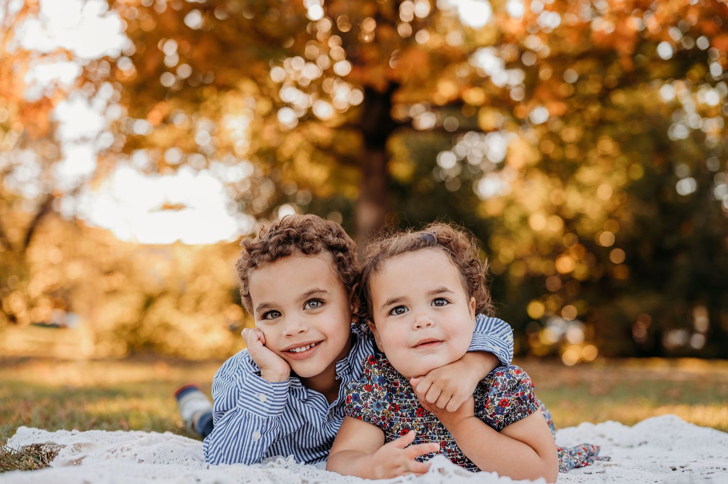 Two children lying on a blanket together outdoors during golden autumn hour with colorful fall foliage in background at a park in Summit.