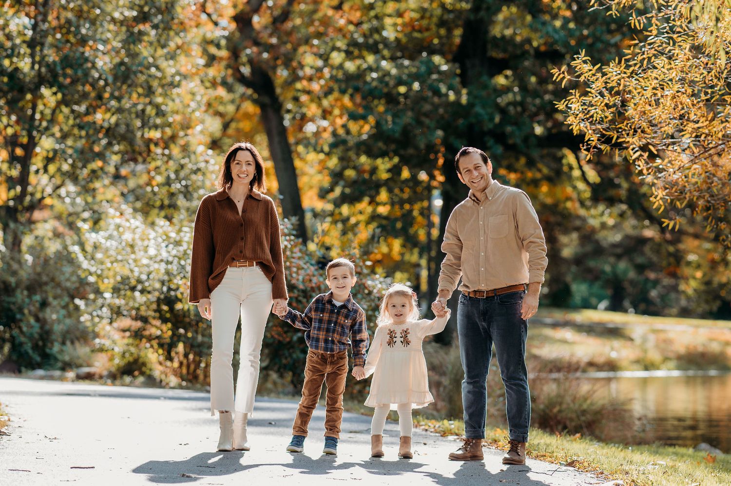 A family of four poses together on a sunny autumn day by a lake with yellow trees in the background at Yantacaw Brook Park in Montclair.