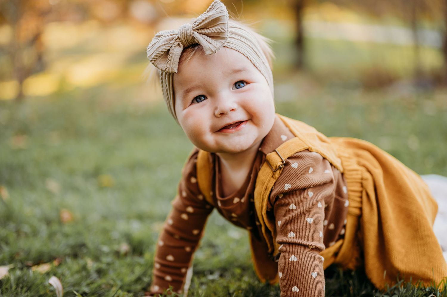 A small child in a mustard yellow outfit and bow headband crawls on grass during autumn.