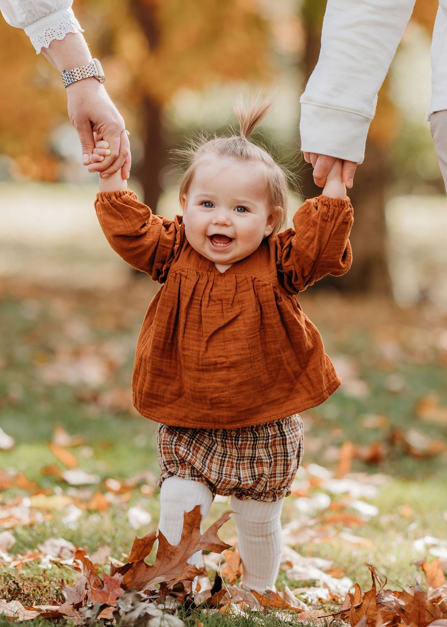Small child in a rust-colored dress and plaid bottoms plays in autumn leaves while holding hands with an adult.