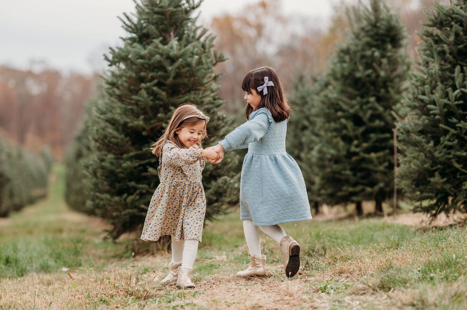 Two young children dance together at a Christmas tree farm on an autumn day, wearing festive dresses.