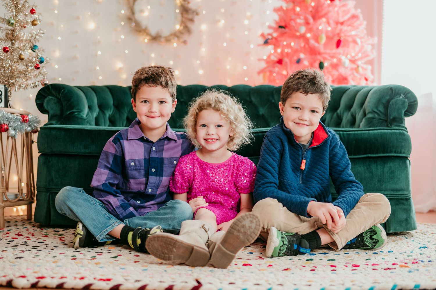 Three children sit together on a green velvet couch with retro vintage Christmas decorations and trees in the background.