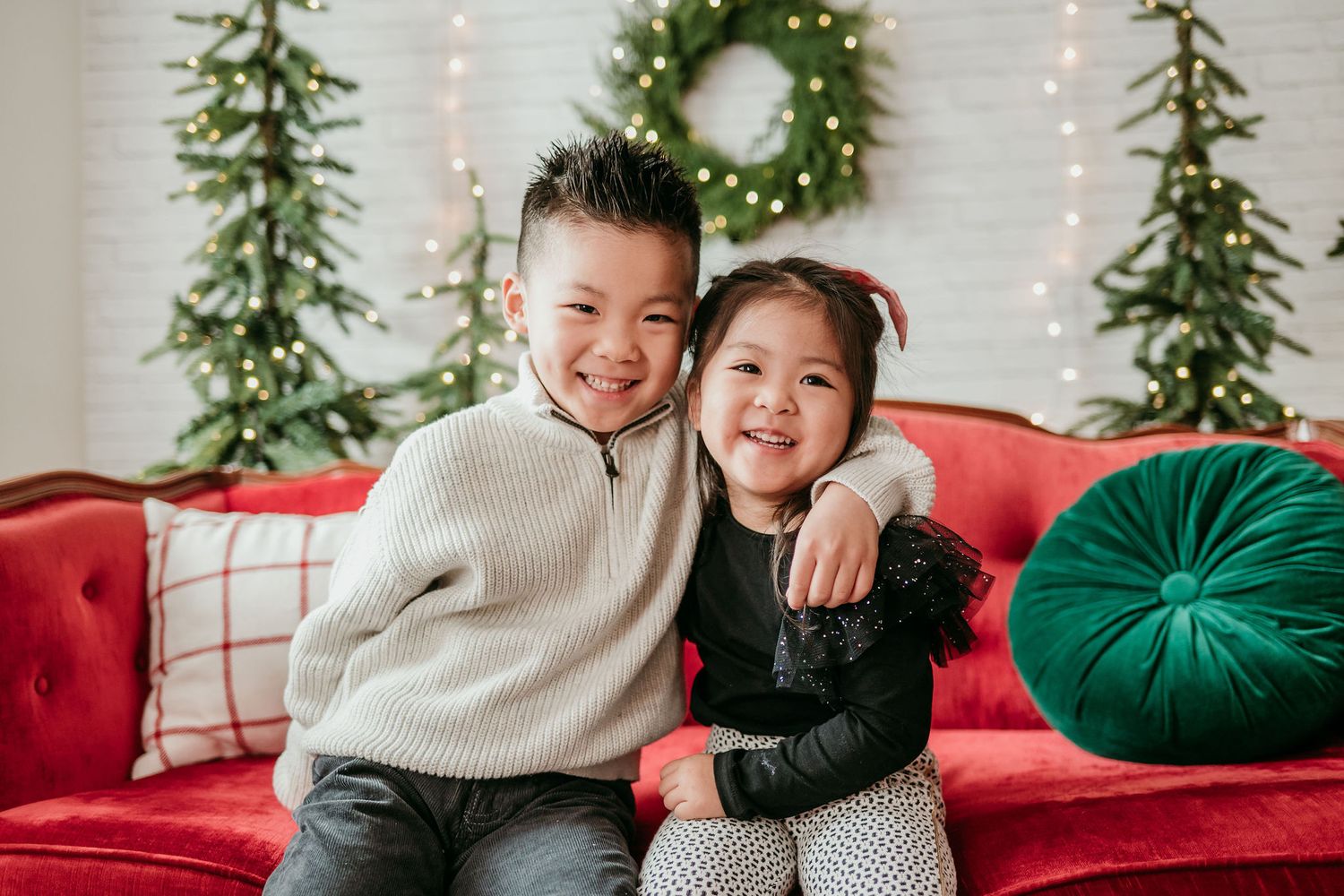 Two children hugging and smiling on a red couch with Christmas decor and greenery in the background.