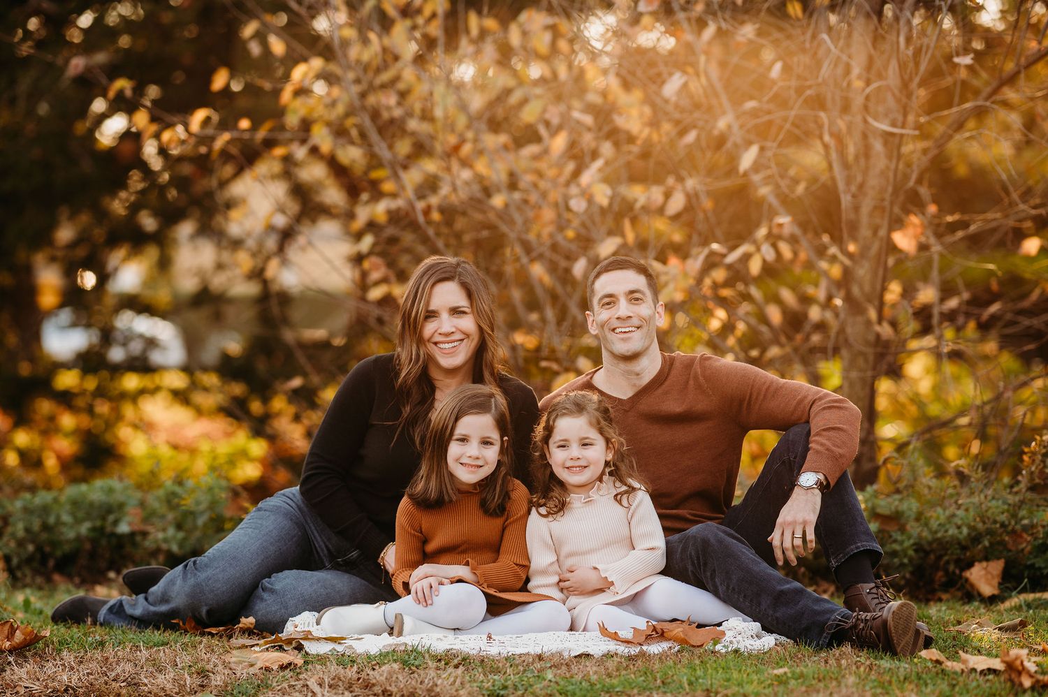 Family portrait outdoors on a blanket surrounded by golden autumn foliage in warm evening light.