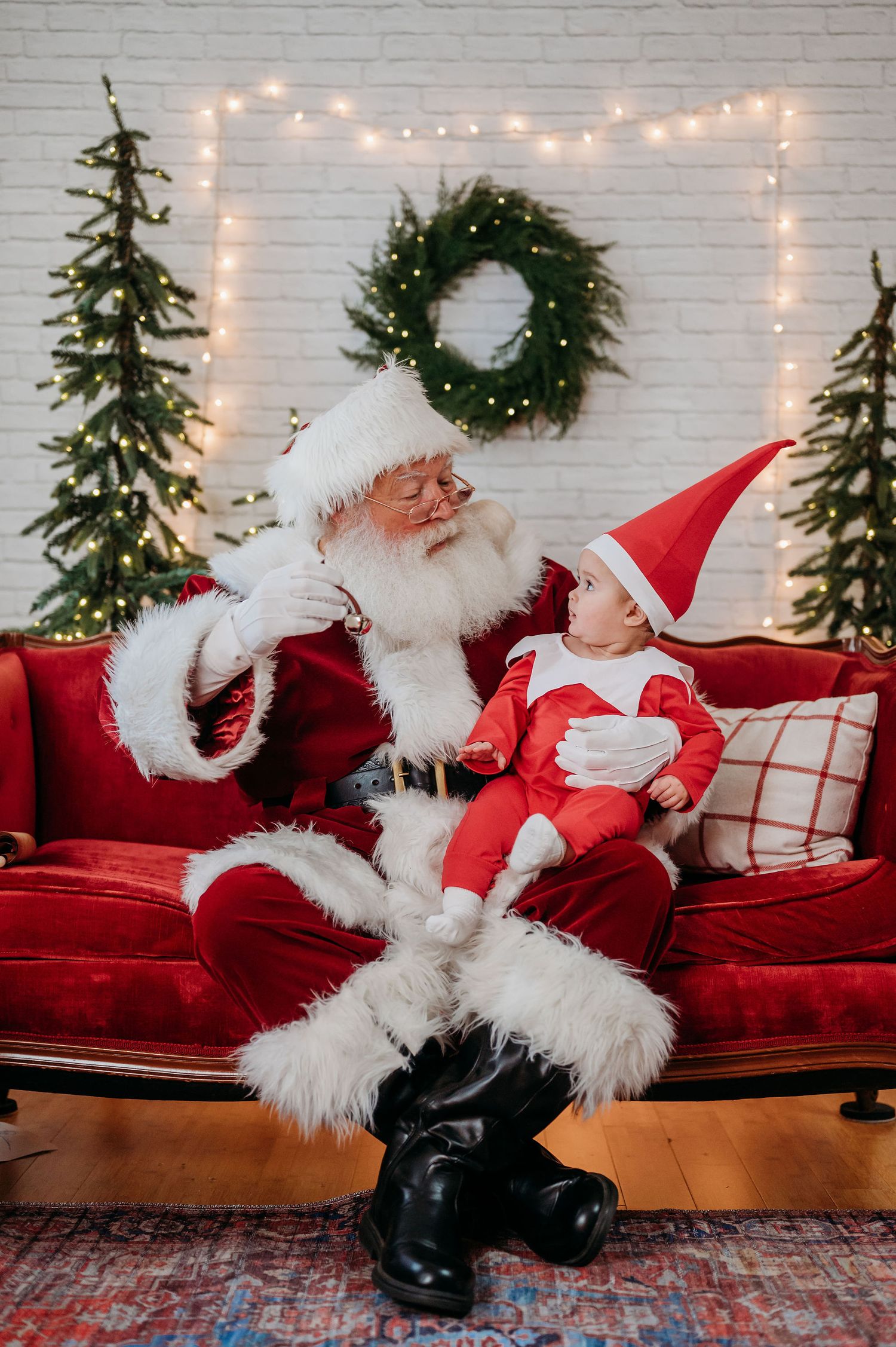 A festive Christmas scene with decorative lighting, evergreen trees and wreath on white brick wall with red velvet sofa. Baby elf with Santa