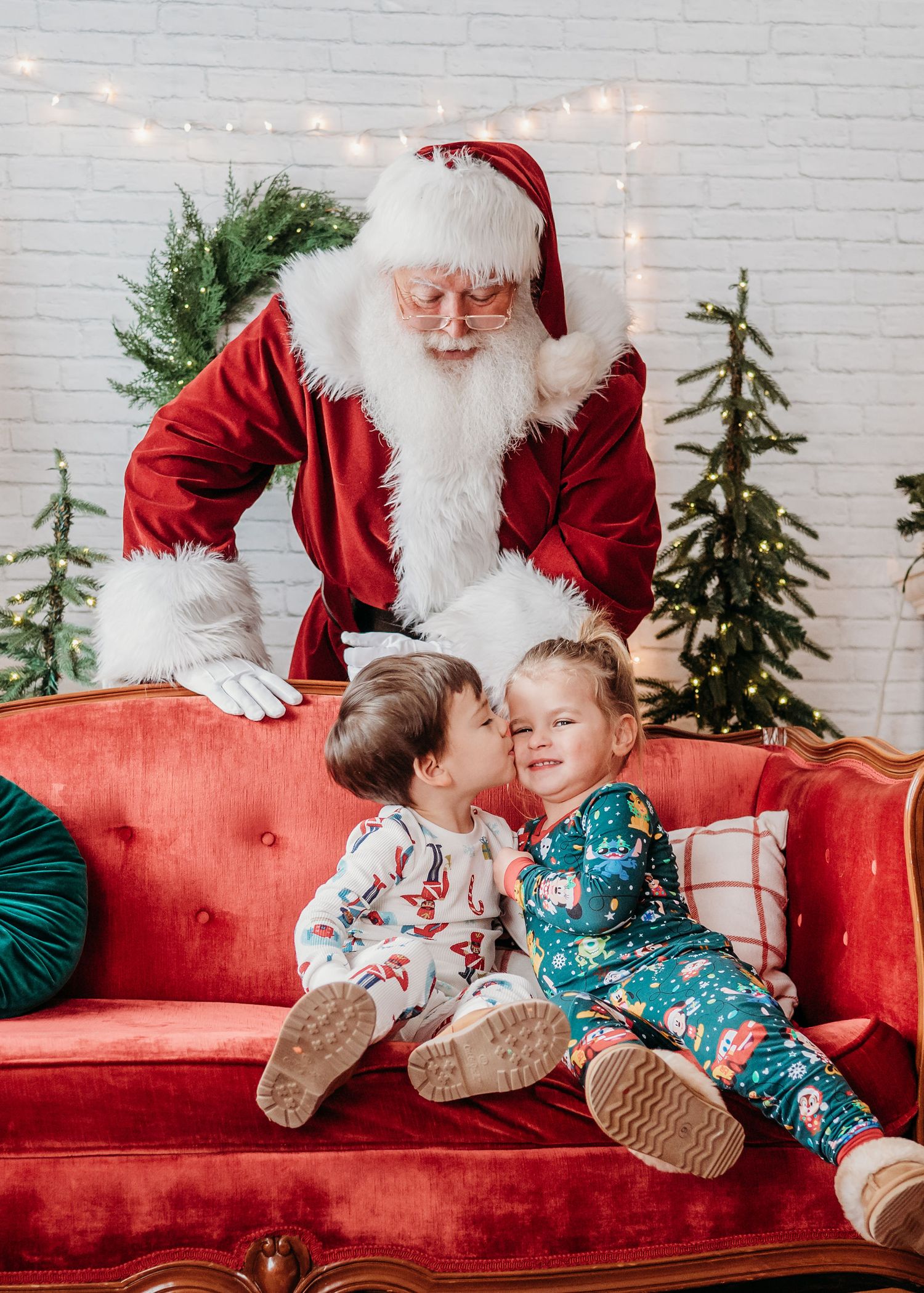 Two children in holiday pajamas sitting on a red velvet couch with festive Christmas decorations kiss as Santa looks down at them.