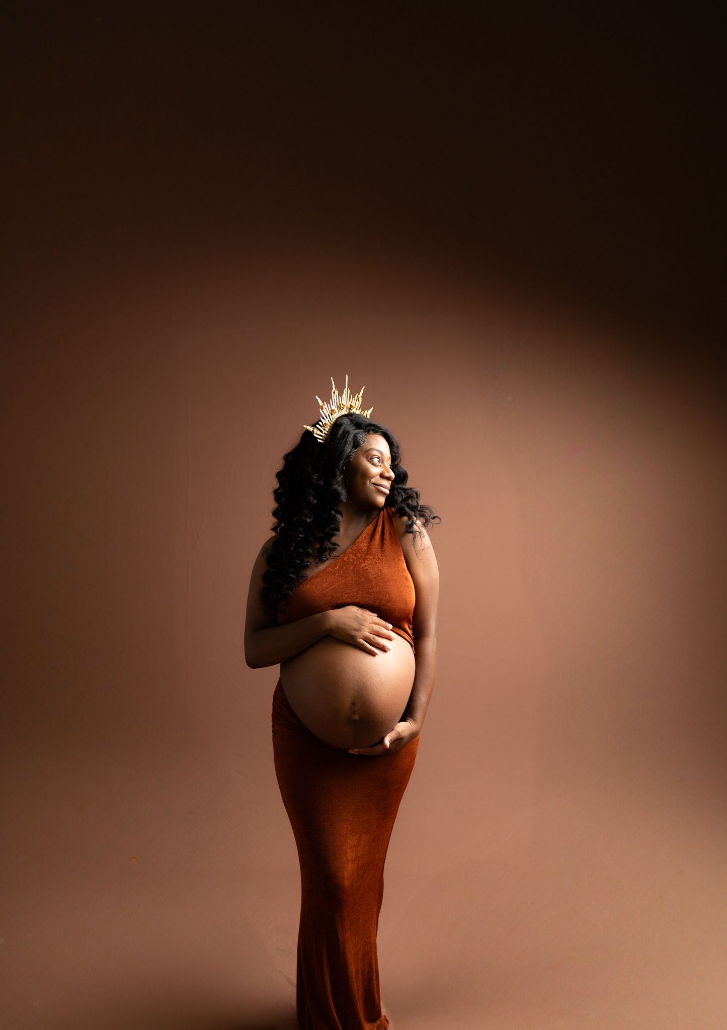 Expecting mother in a rust-colored velvet gown poses for a maternity portrait against a neutral backdrop.