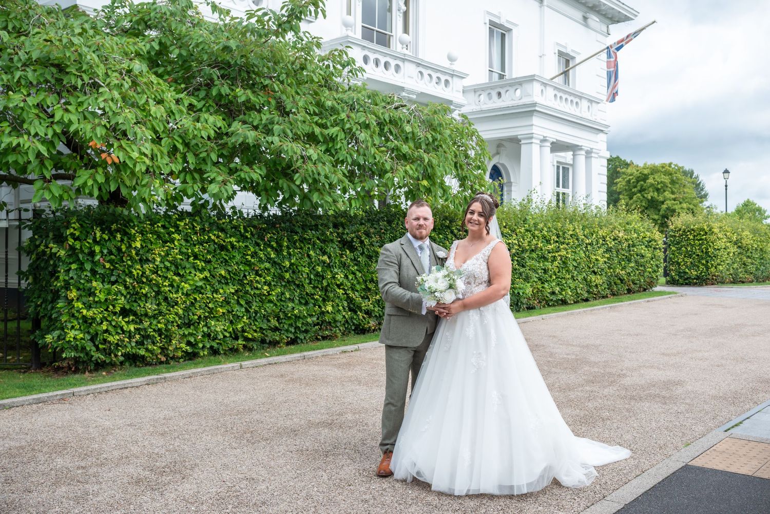 A couple in wedding attire walk together along a scenic outdoor path surrounded by lush green hedges and architecture.