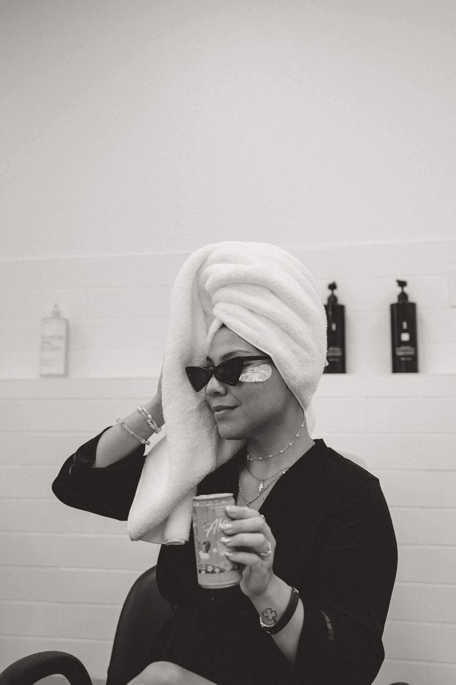 Black and white photo showing figure in sunglasses and white hair towel holding coffee cup in relaxed spa-like setting.