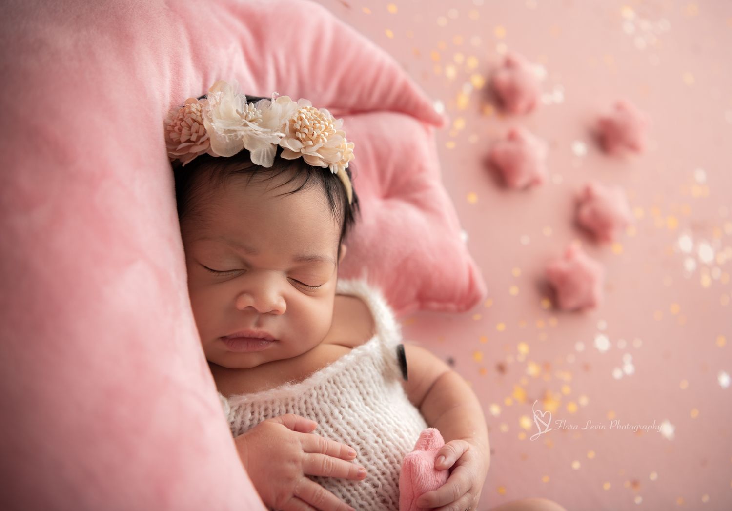 Sleeping newborn on pink moon wearing white knit outfit and floral headband with soft sparkle overlay effect. On a pink backdrop with stars