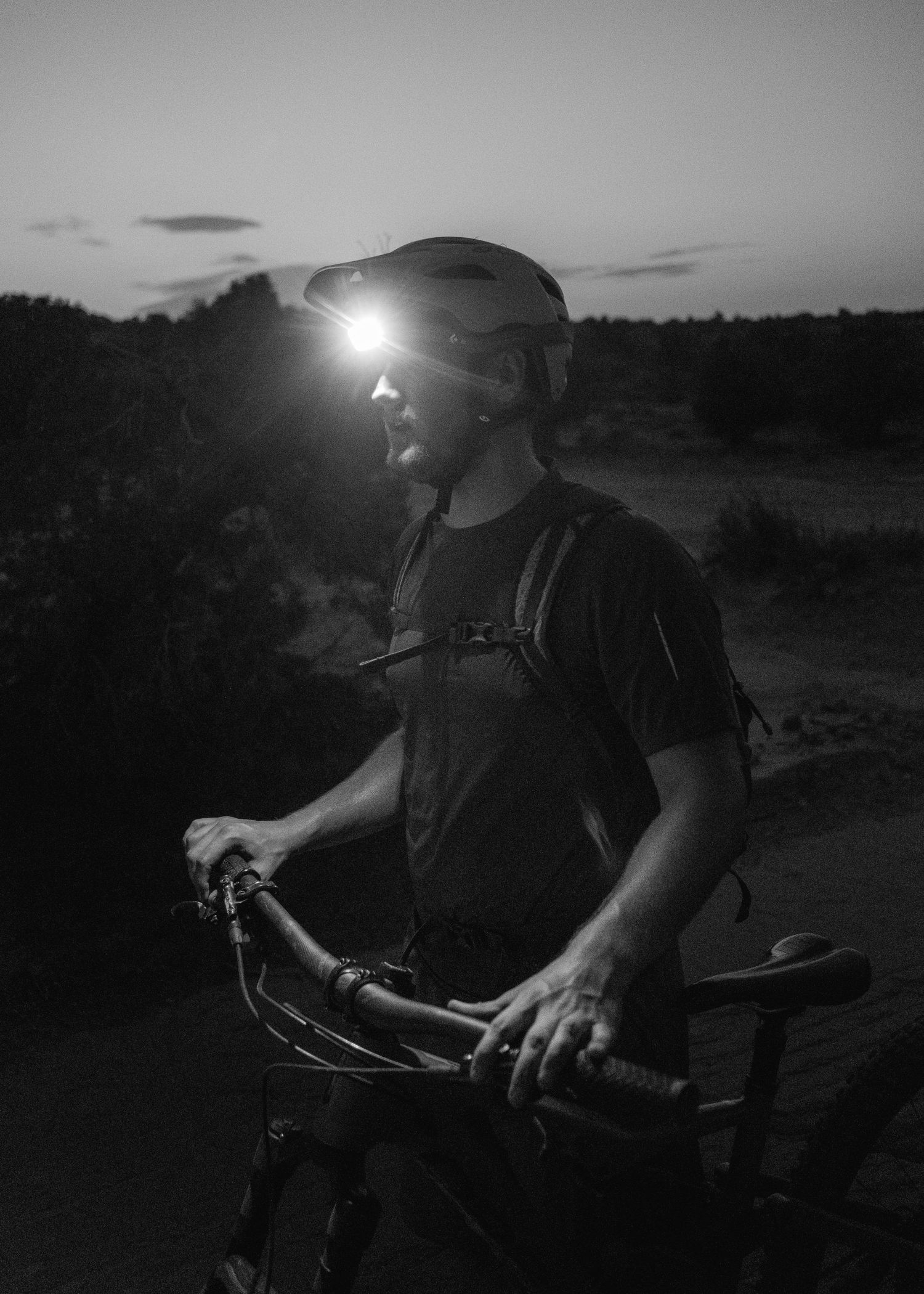 Mountain biker with headlamp silhouetted against dusky sky in dramatic black and white photo.