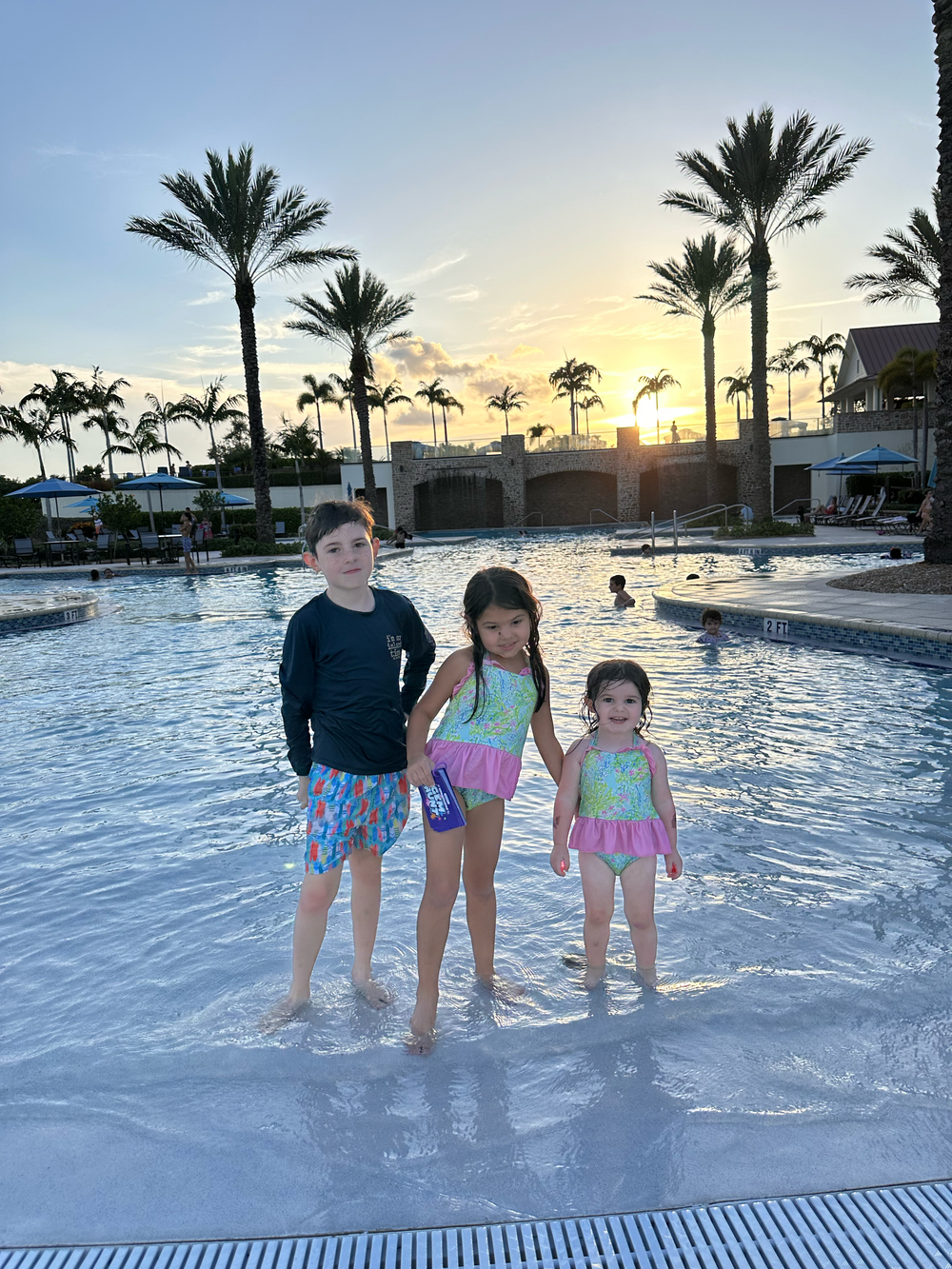 People standing in a swimming pool at sunset with palm trees silhouetted against the orange sky.