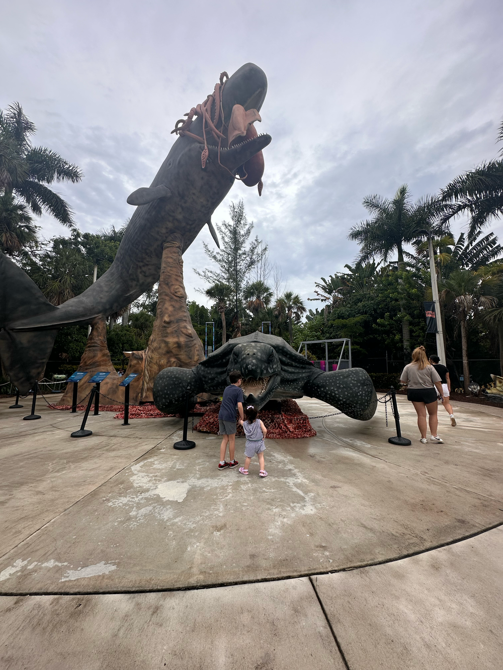 Large decorative dinosaur statues displayed in an outdoor entertainment area with palm trees.