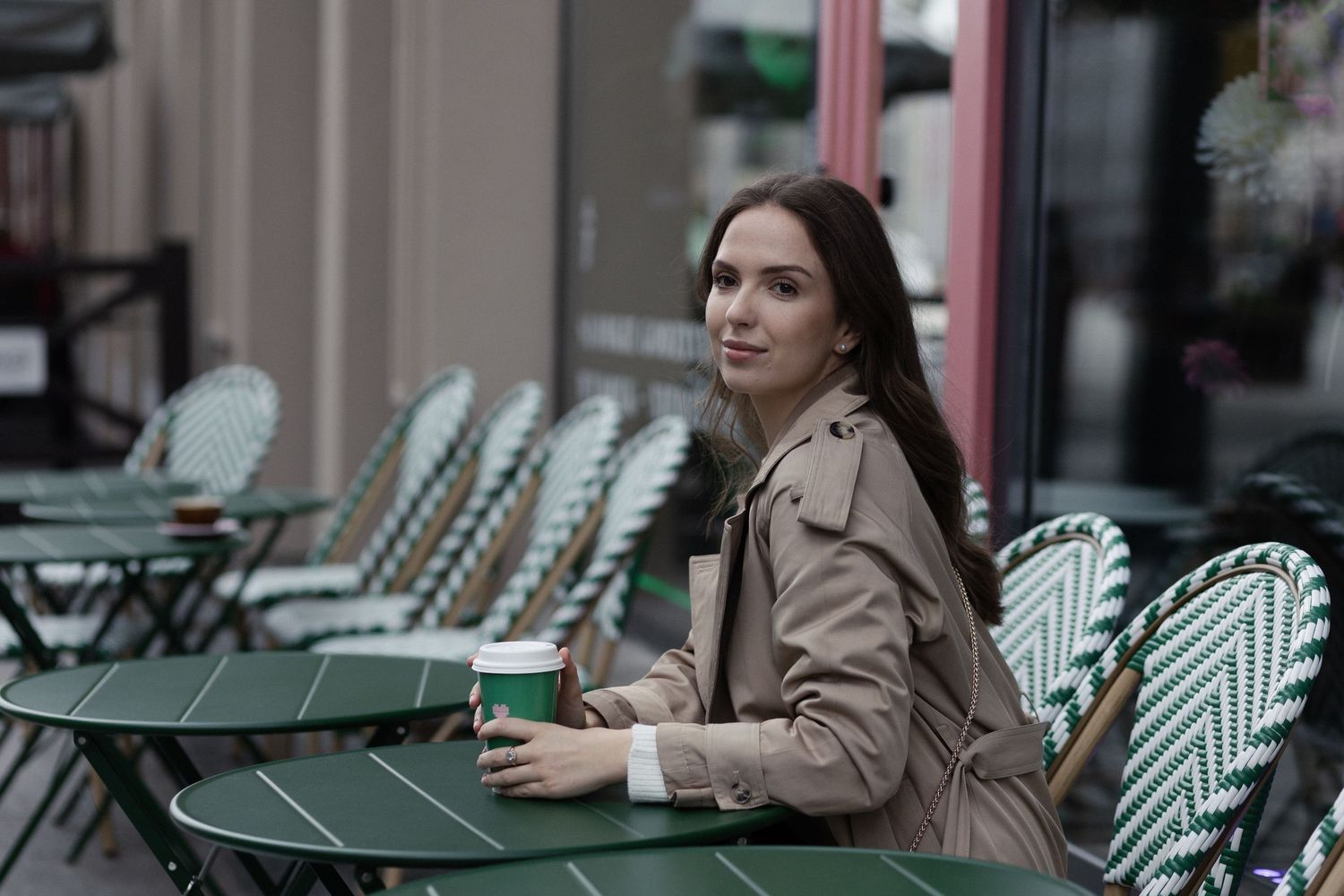 Someone in a beige trench coat sits alone at an outdoor Parisian-style cafe with green bistro chairs and tables.