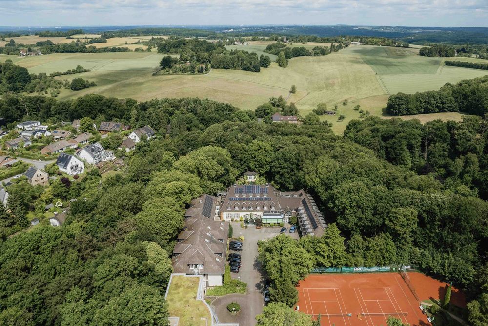 Aerial view of a countryside estate with tennis courts and buildings surrounded by lush green forests.