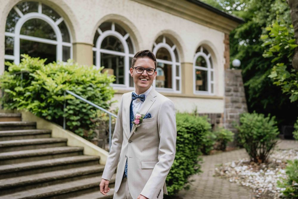 Formal portrait of a person in a light colored suit with boutonniere against stone building backdrop.