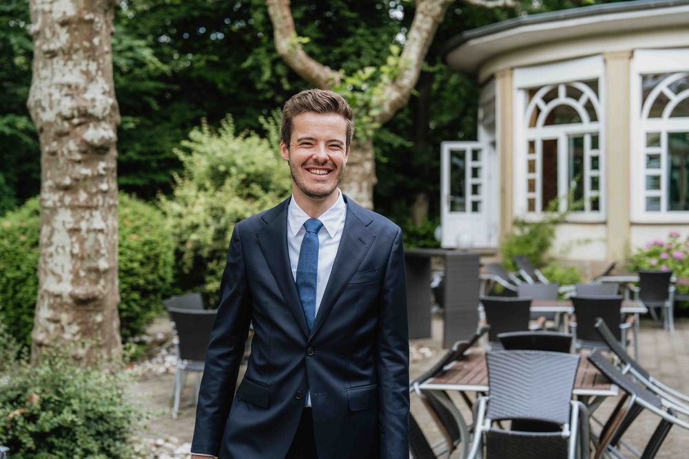 Person in navy suit smiling outdoors near classic architecture and garden furniture.