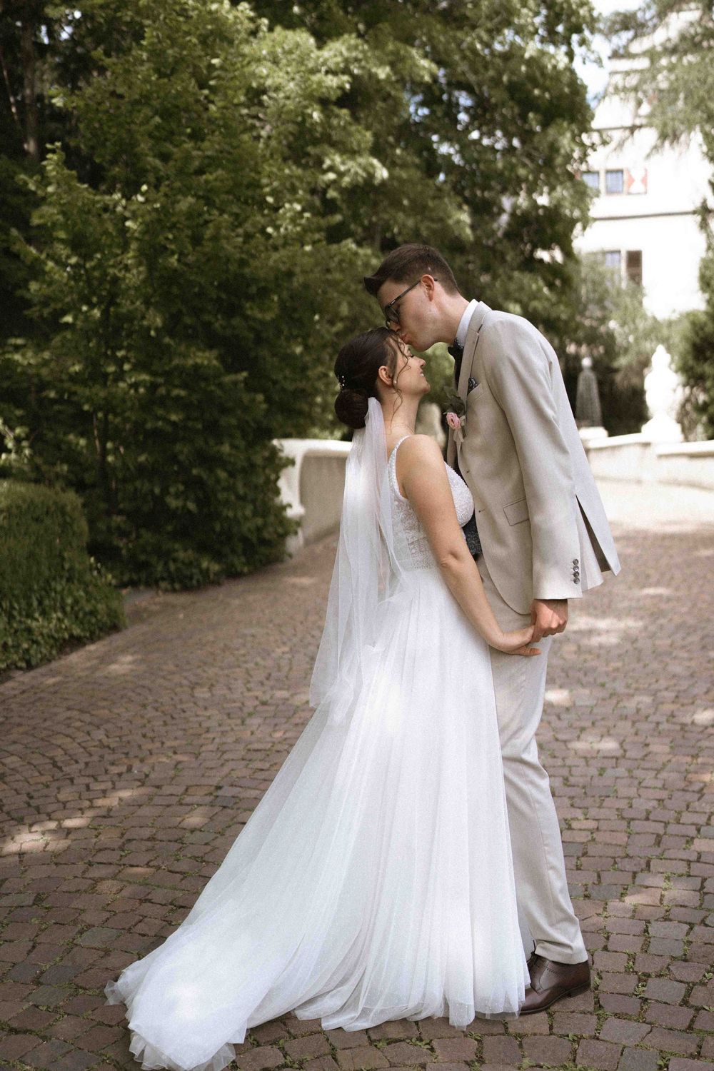 Wedding couple sharing a kiss on a cobblestone path surrounded by greenery.