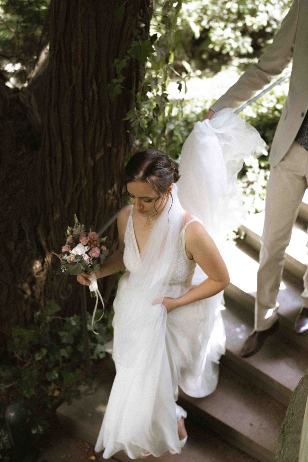 Bride in flowing white dress holds bouquet while descending garden steps.