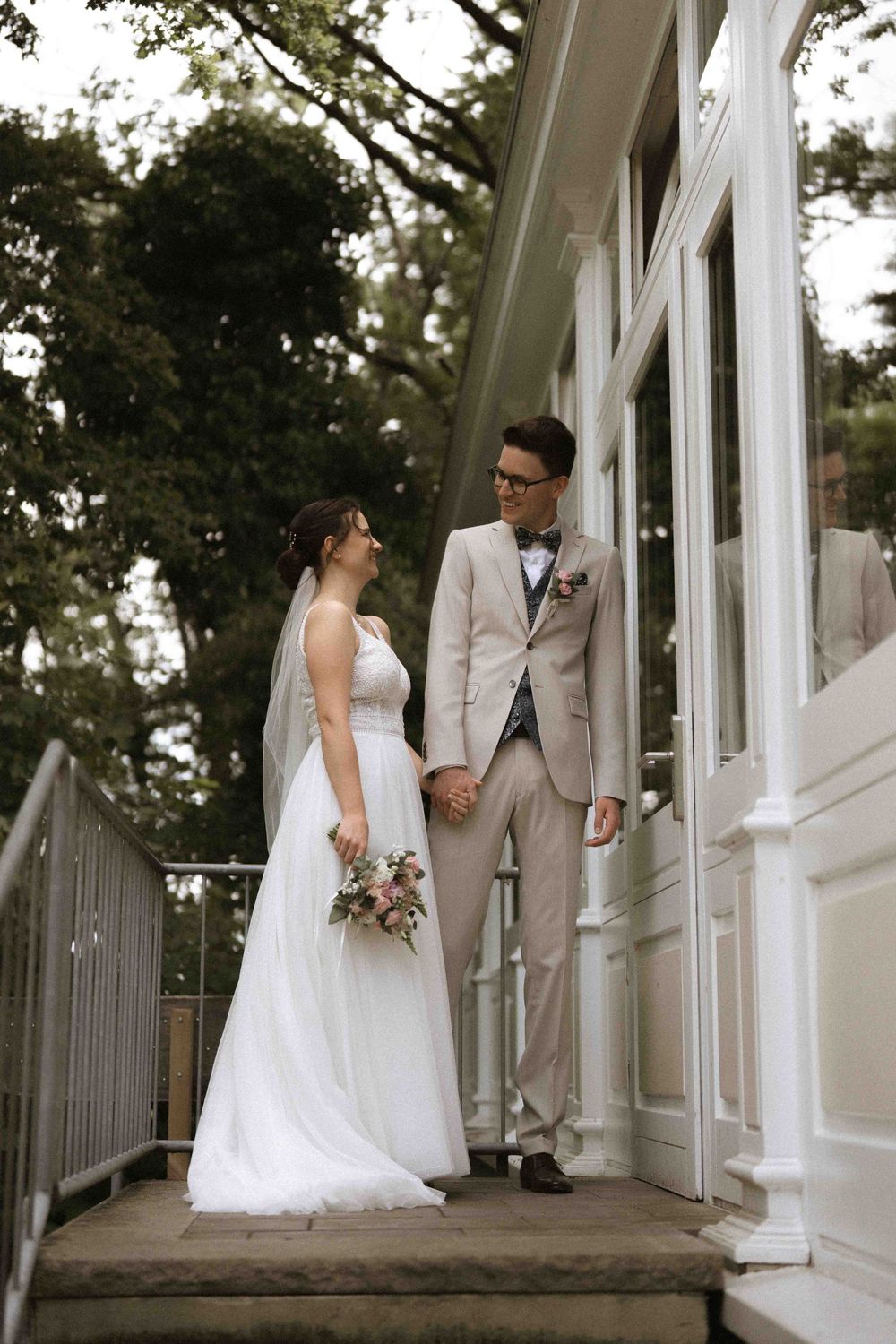 Couple in wedding attire standing together on white porch steps.