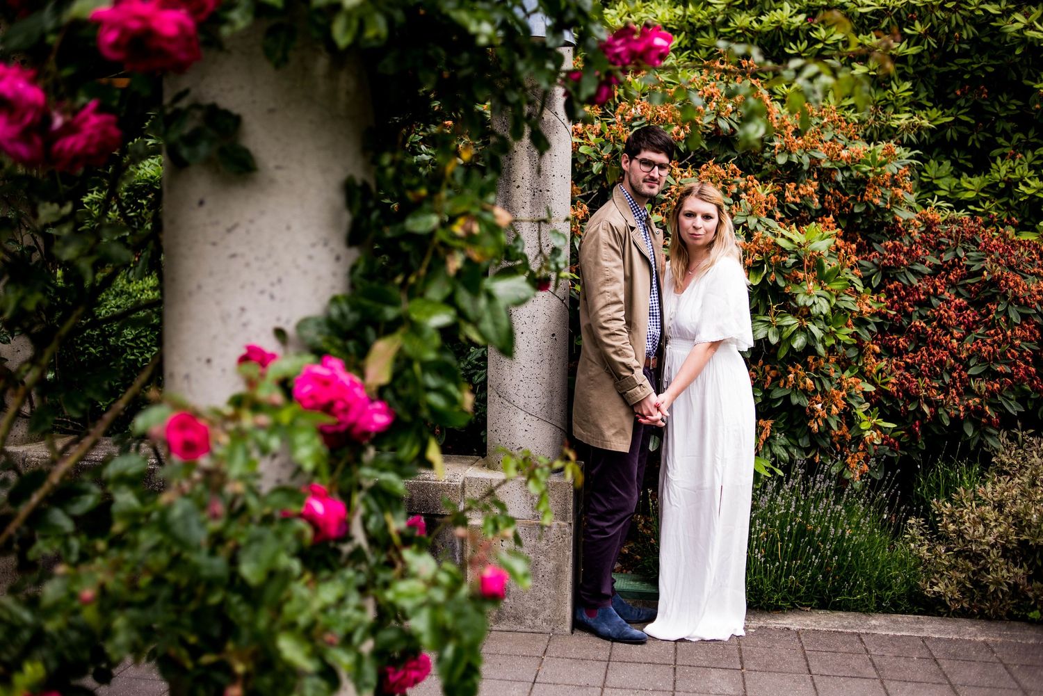 Two people pose together in a lush garden setting with vibrant pink roses and greenery in the background.