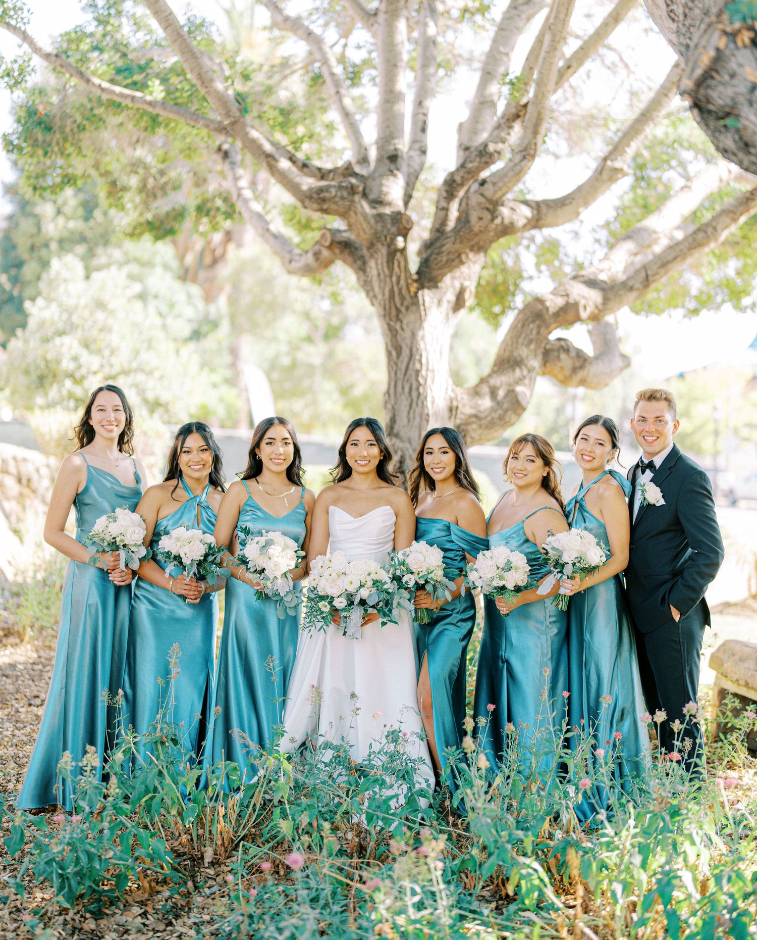 Wedding party in turquoise dresses poses together under a large tree.