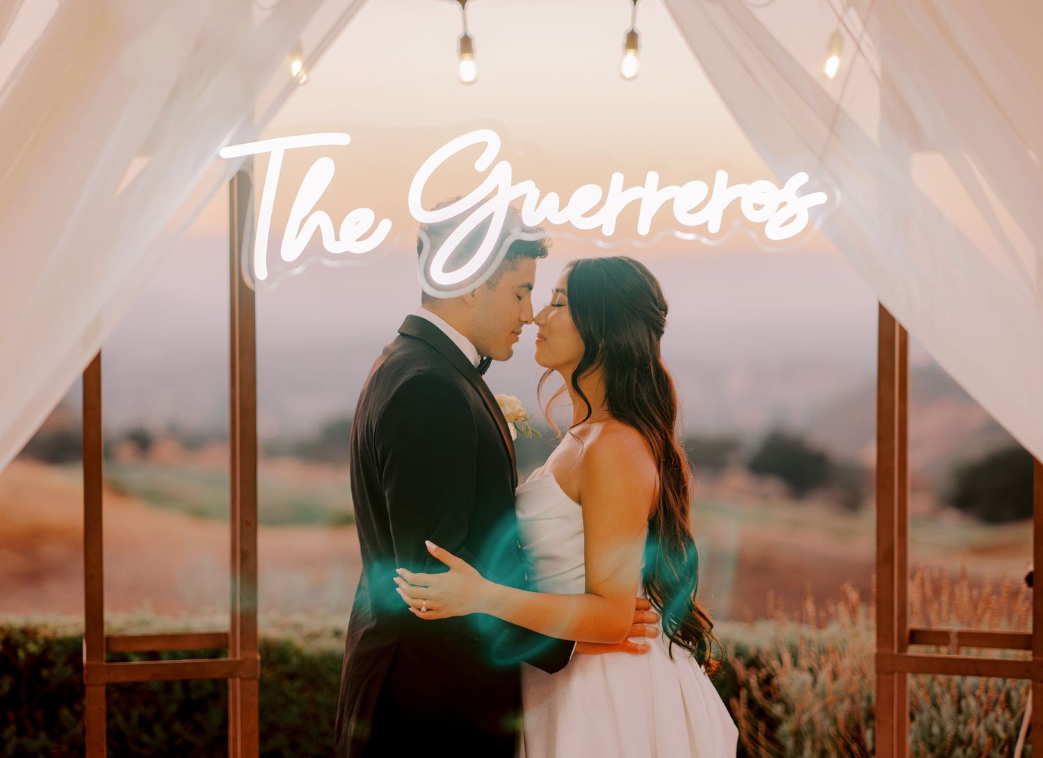 Intimate wedding portrait under a white canopy with neon sign and sunset backdrop.