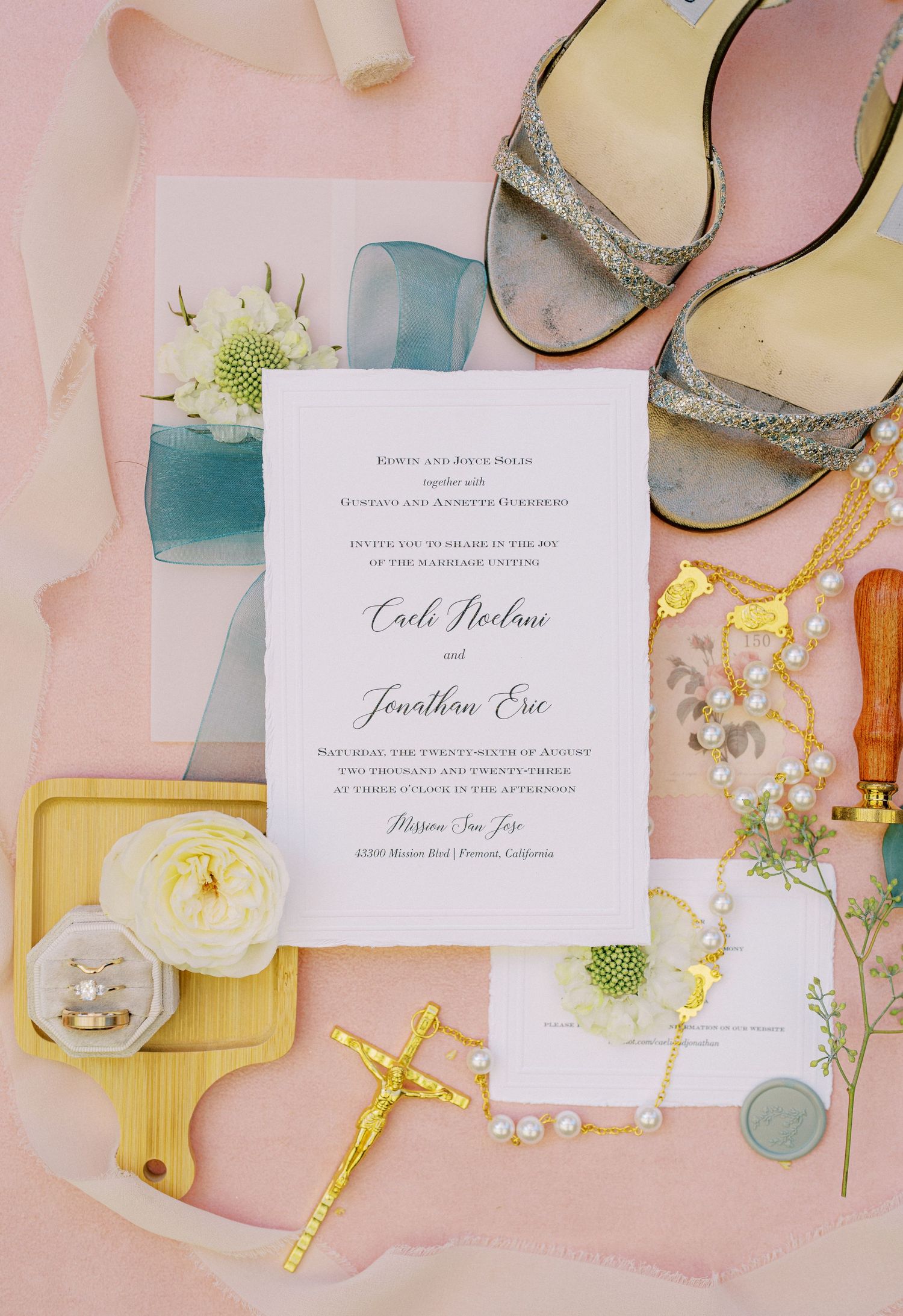 Wedding invitation and accessories including silver shoes and religious items on pink background.