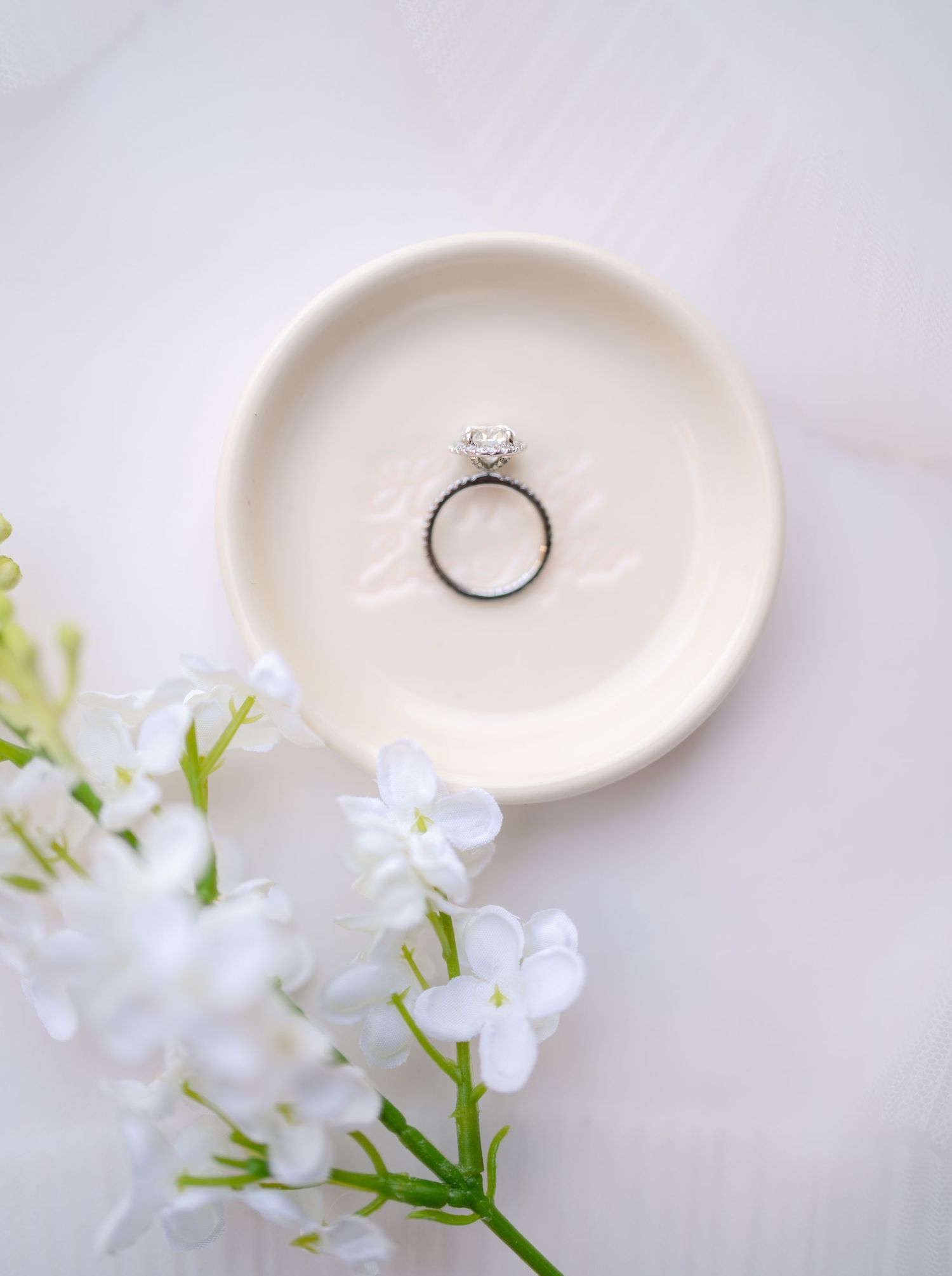 Diamond engagement ring displayed on white ceramic plate with delicate white flowers.
