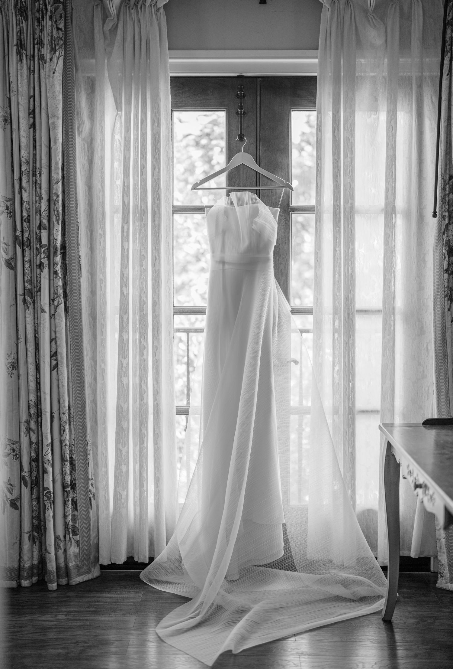 Wedding dress hanging in window with flowing curtains in black and white photography.