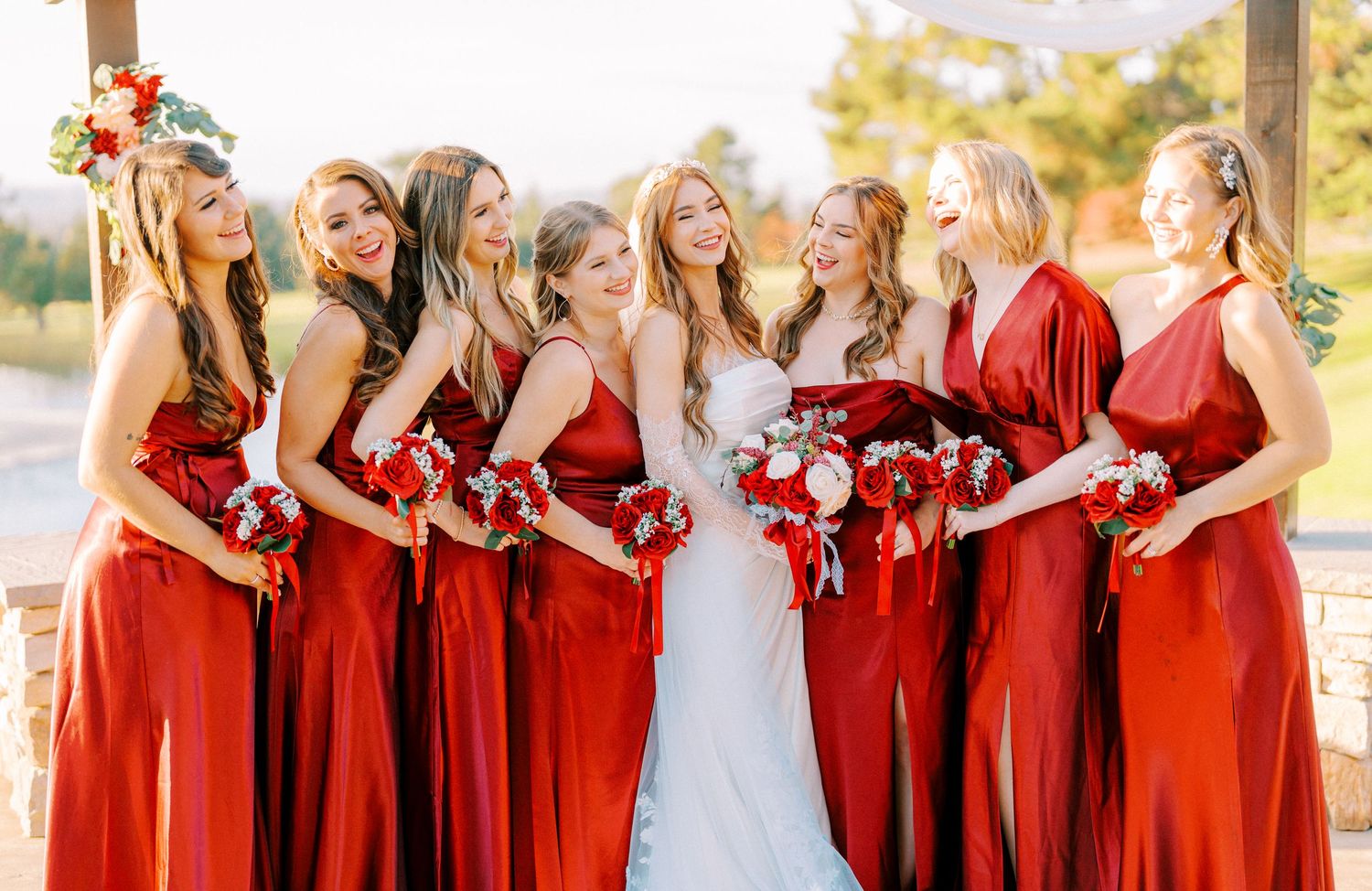 Wedding party in vibrant red bridesmaid dresses pose together outdoors in warm sunlight.