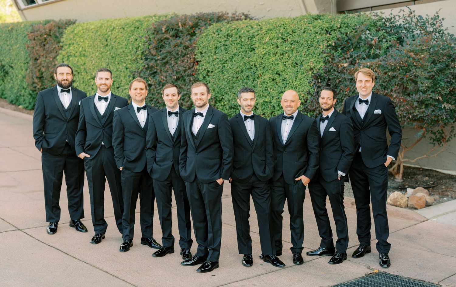 Groomsmen in matching black tuxedos and bow ties lined up outdoors for a formal wedding photo.