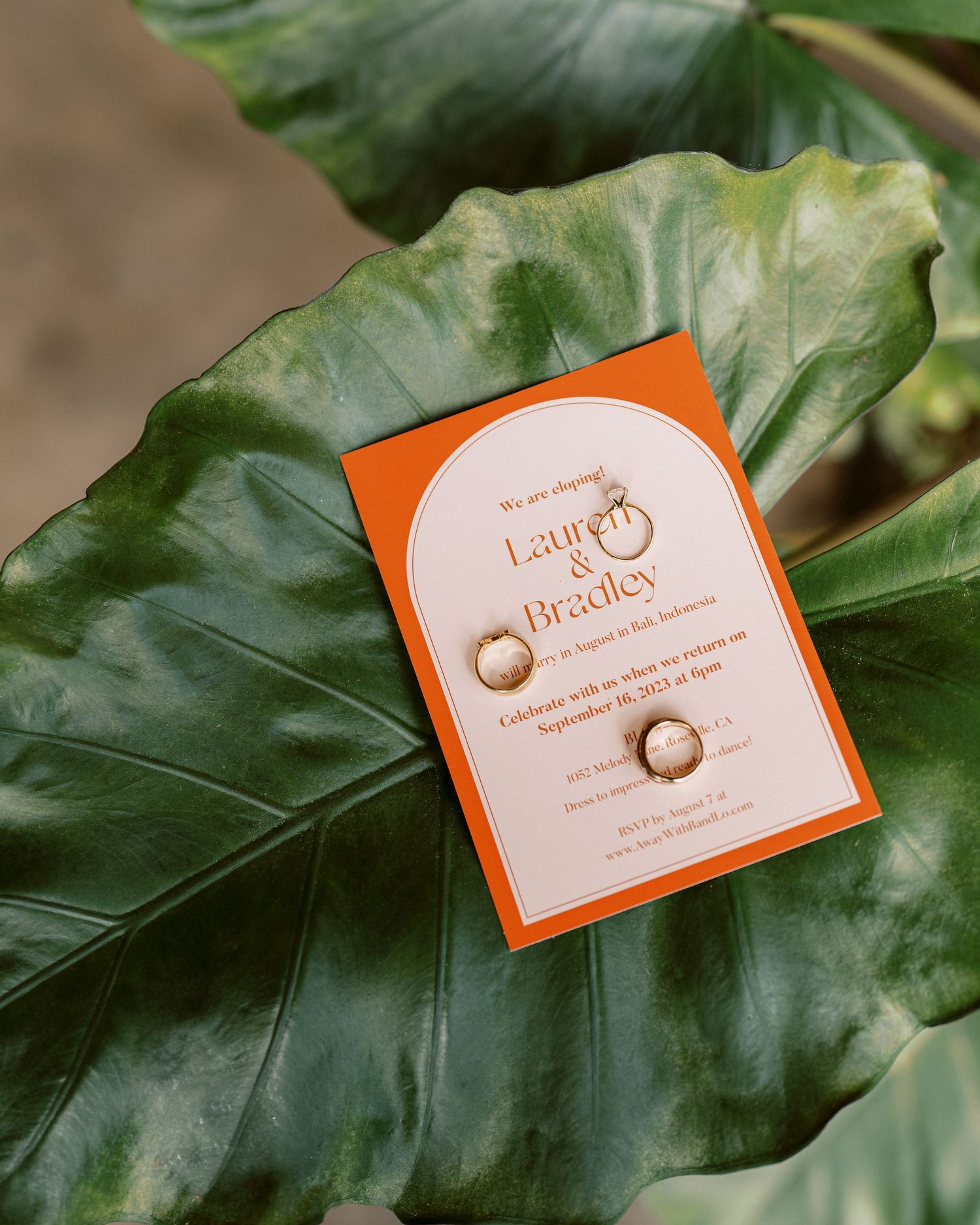 Orange and white wedding invitation displayed on large green tropical leaf.
