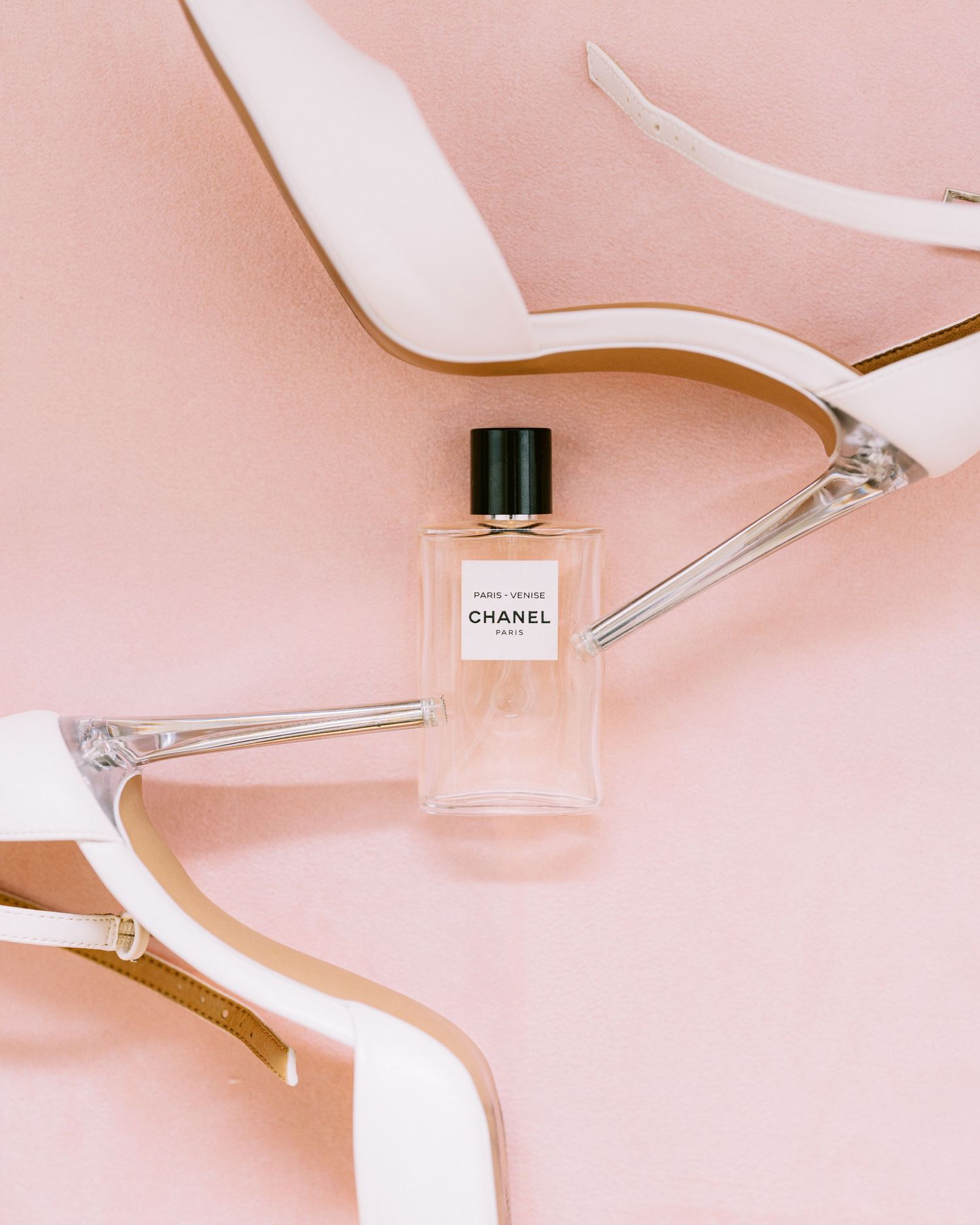 White bridal heels and Chanel perfume bottle styled on pink background.