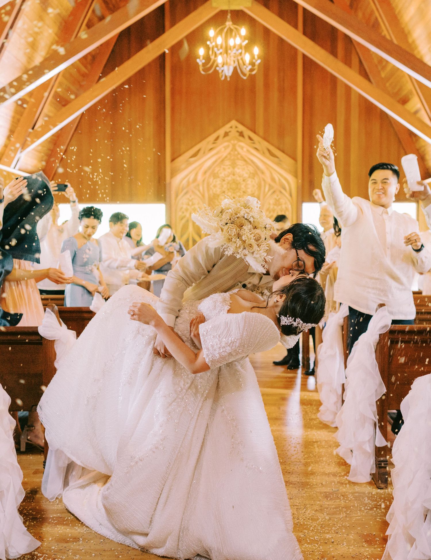 Newlyweds share a romantic dip during their first dance in a wood-paneled chapel with hanging lights.