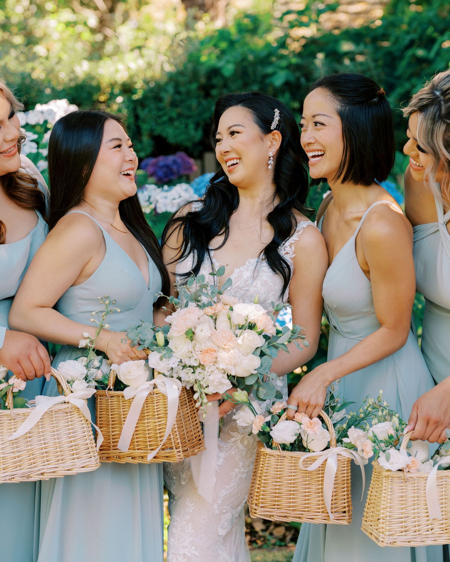 Bridesmaids in light blue dresses carry wicker baskets with floral arrangements while sharing a joyful moment.