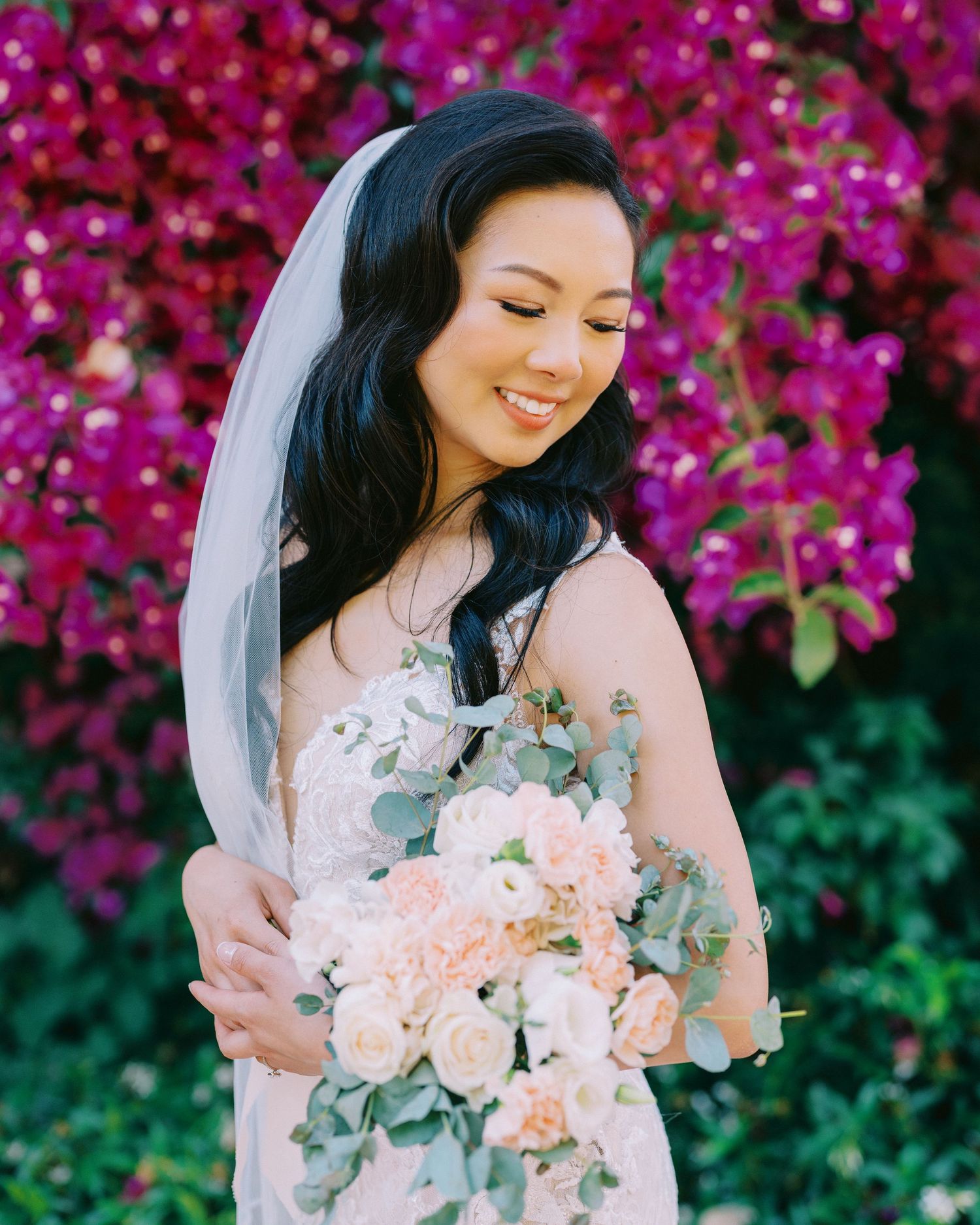 A bride holds a bouquet of blush roses against a vibrant magenta bougainvillea background.