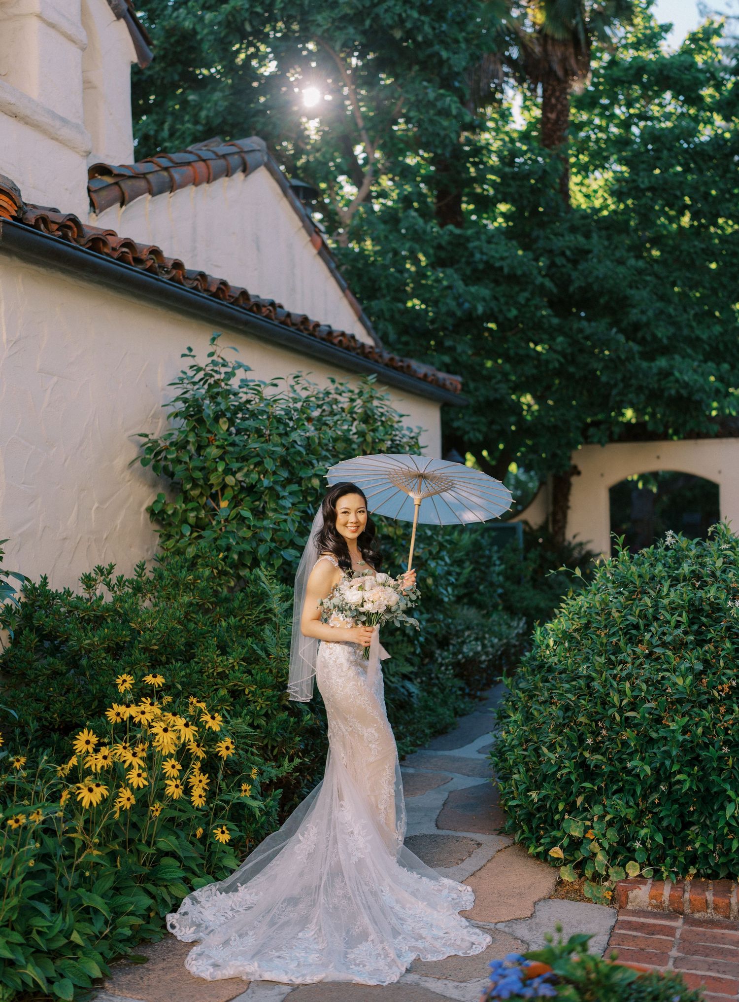 A bride in a lace mermaid dress poses with a parasol in a garden setting with yellow flowers and sunlight.