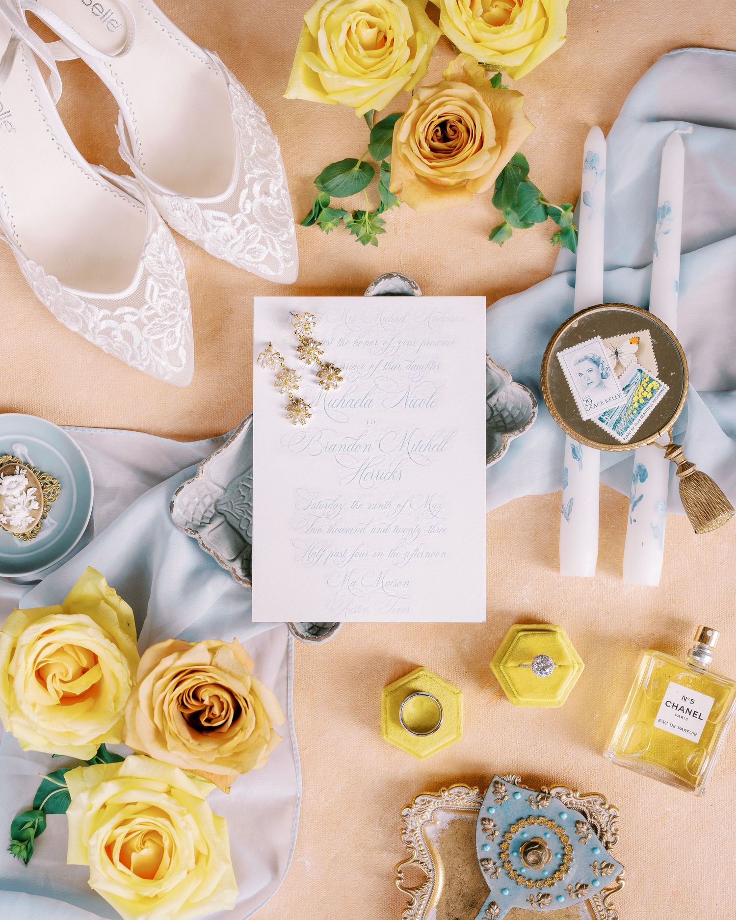 Wedding flatlay with lace shoes, yellow roses, perfume bottle and invitation on peach background.