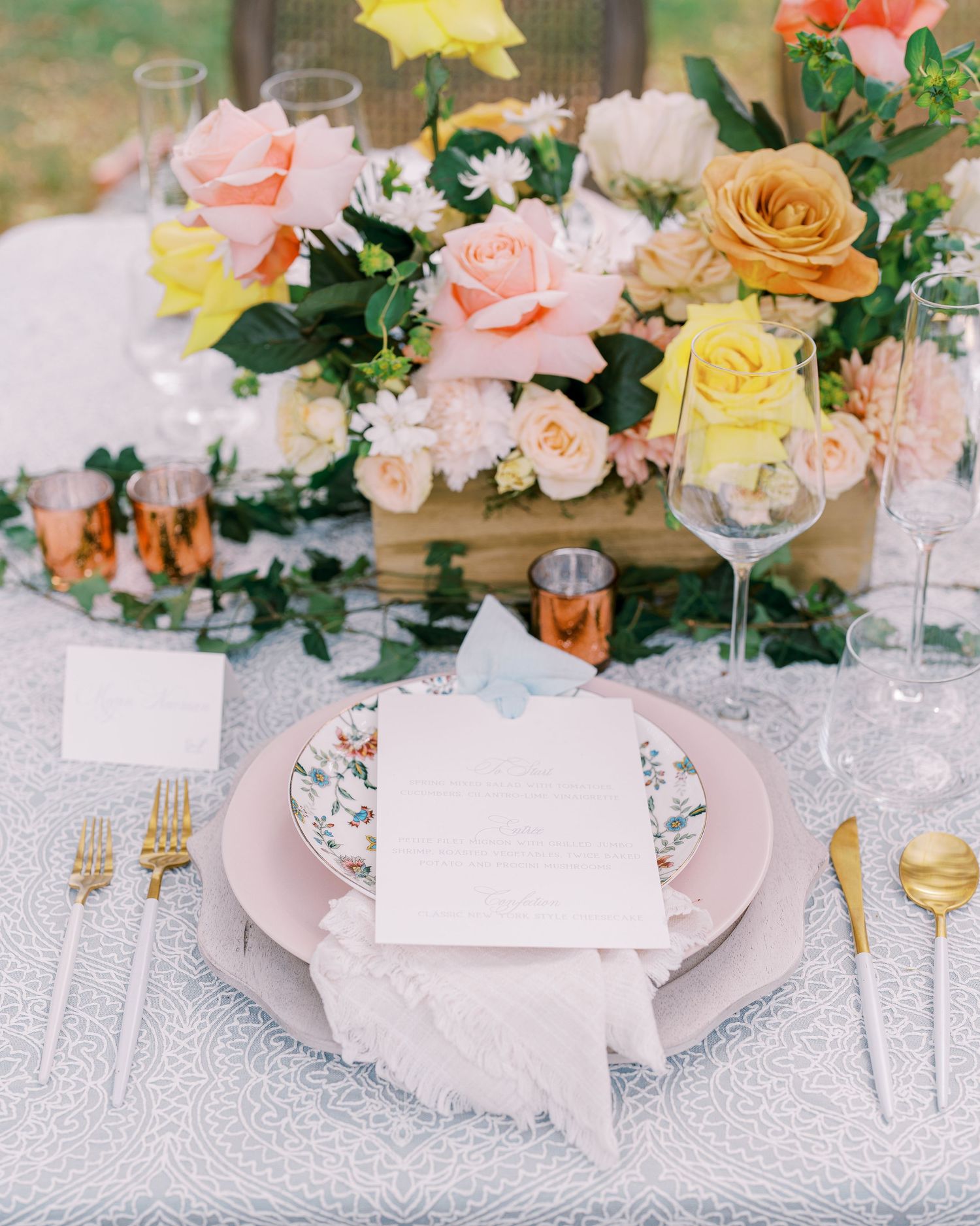 Romantic place setting with pink and yellow floral centerpiece and gold flatware on lace tablecloth.