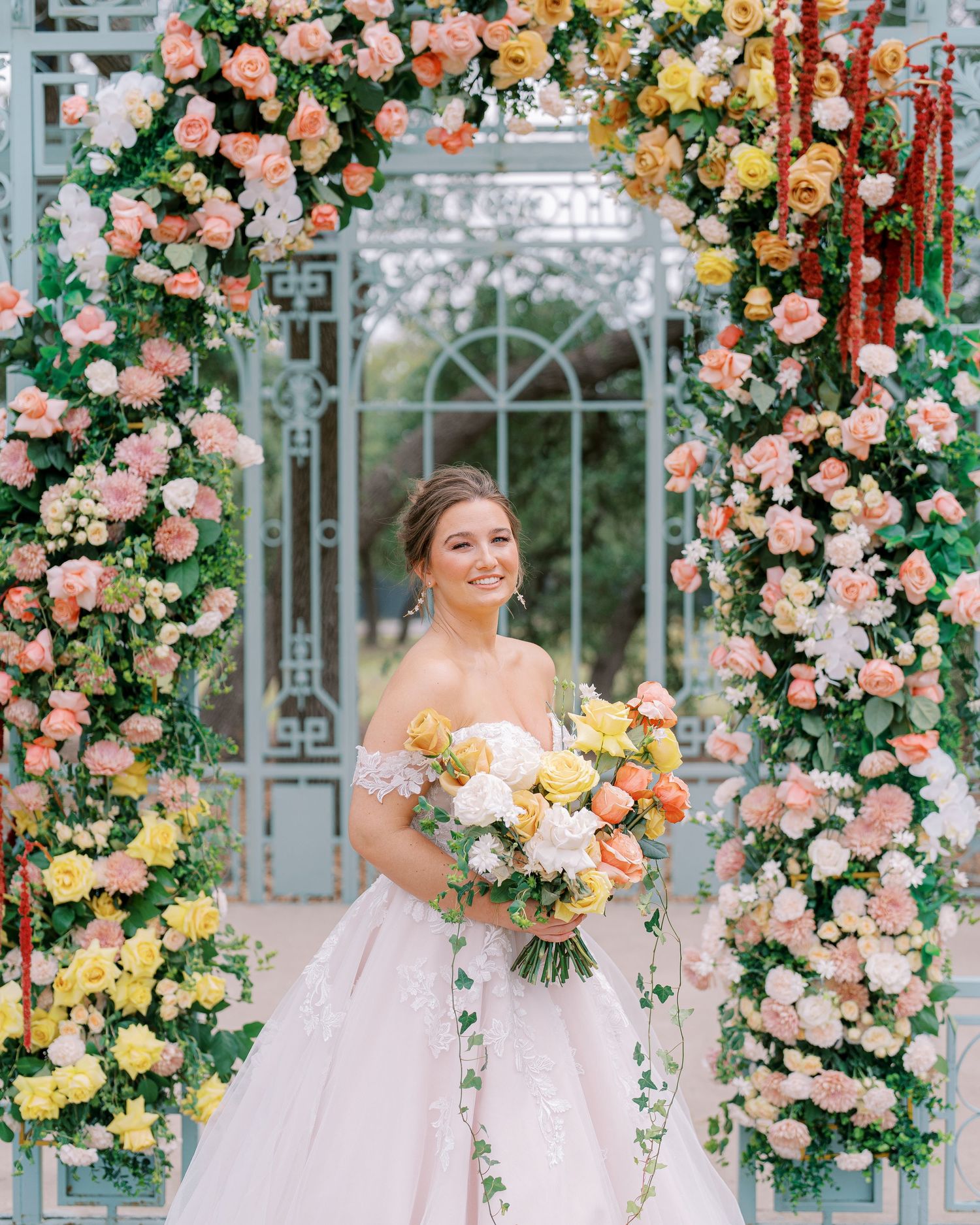 Beautiful floral arch with pink and yellow roses frames an ornate glass greenhouse.