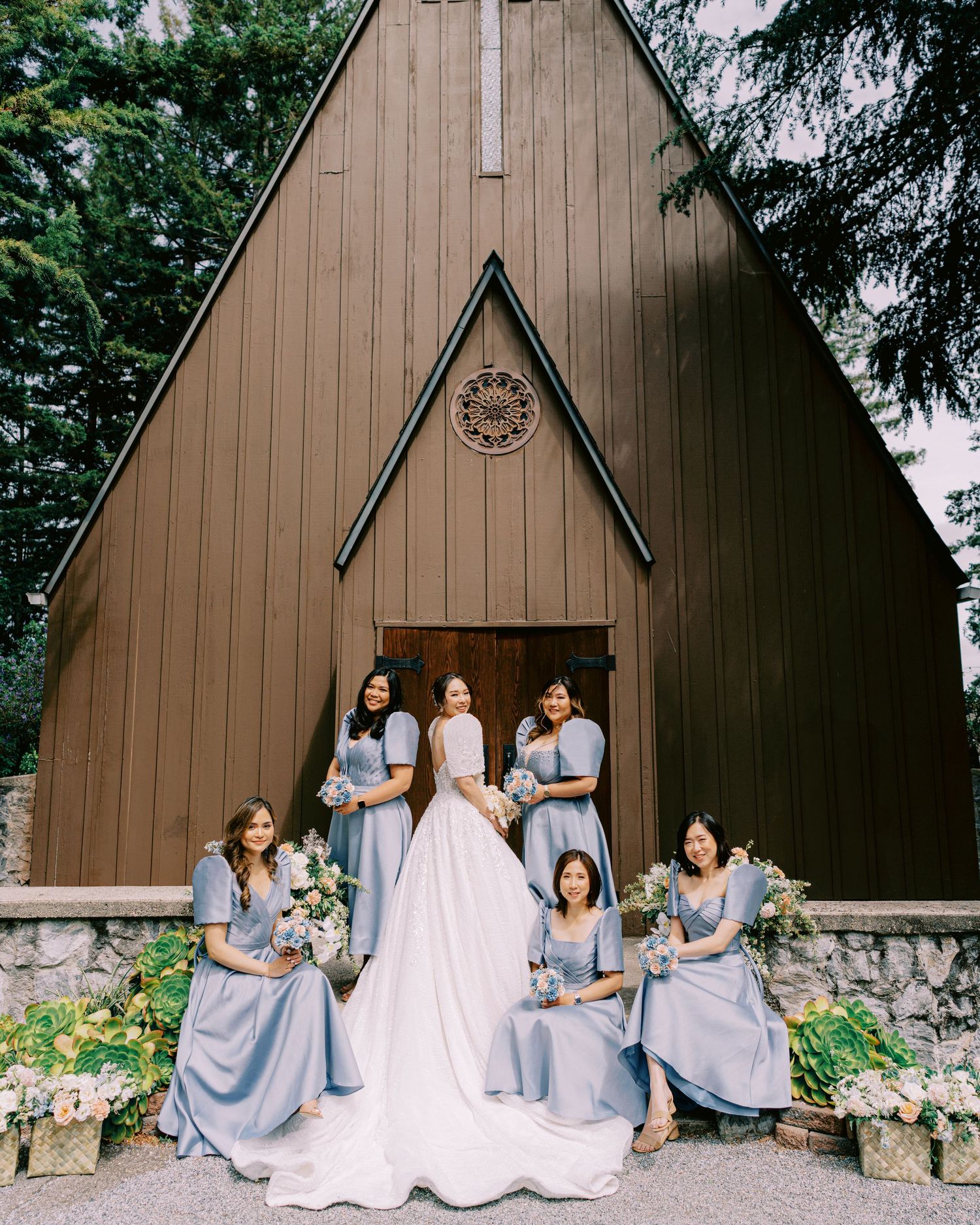 Wedding party poses on steps of a rustic brown A-frame chapel with light blue bridesmaid dresses.