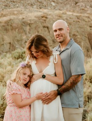 Expectant parents and young girl in pink dress pose together against rocky desert backdrop at sunset.