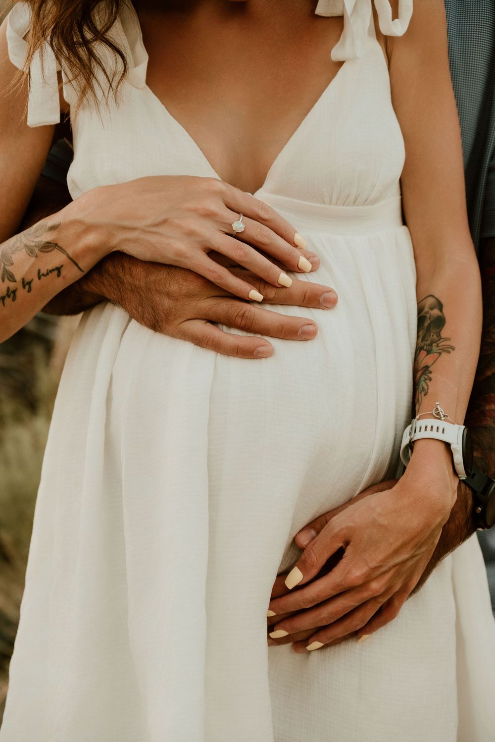 Close up of hands with tattoos and a silver watch embracing a white maternity dress with bow straps.