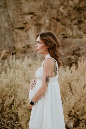 An expectant mother in a flowing white dress poses in profile against golden grasses and rocky terrain.